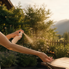 Frau sprüht Mückenspray auf ihren Arm auf sonniger Terrasse mit Garten und Bergen im Hintergrund